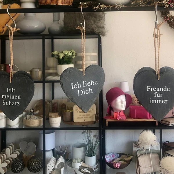 Drei herzförmige Schiefertafeln mit deutschen Sprüchen hängen vor einem Regal mit Wohnaccessoires.