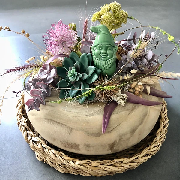 Dekoschale mit Trockenblumen, Sukkulenten und einer grünen Zwergenfigur auf einem geflochtenen Untersetzer.